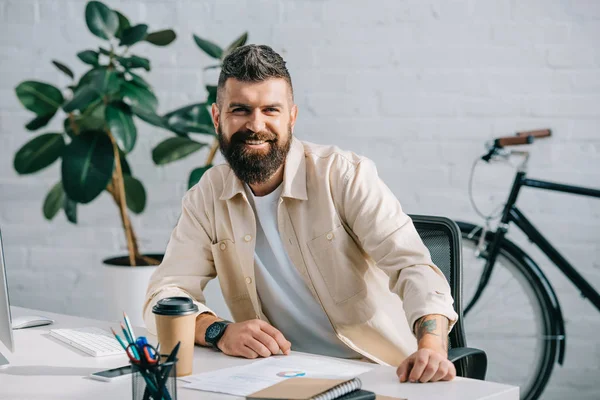 Empresario barbudo sentado en la oficina moderna y sonriendo a la cámara - foto de stock