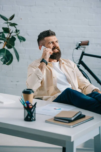 Homem de negócios barbudo sorridente sentado no escritório moderno e falando no smartphone — Fotografia de Stock