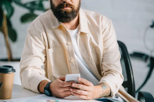 Обрезанный вид бородатого человека, сидящего в офисе и держащего смартфон — стоковое фото