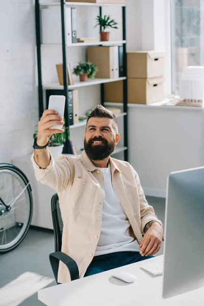 Улыбающийся бородатый бизнесмен делает селфи, сидя в офисном кресле — стоковое фото