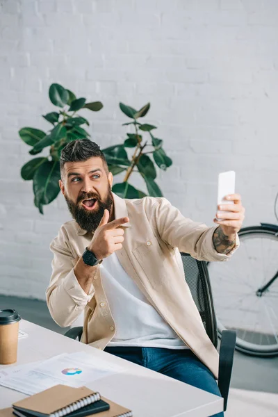 Bearded businessman taking selfie on smartphone and gesturing in modern office — Stock Photo