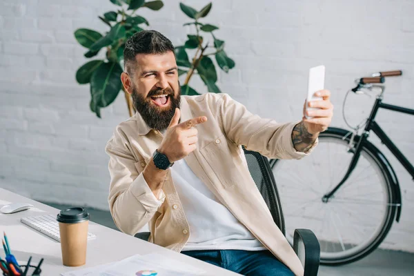 Empresario barbudo apuntando a la cámara mientras toma selfie en silla de oficina - foto de stock