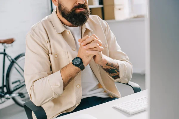 Обрезанный вид бородатого бизнесмена с сжатыми руками, сидящего за компьютерным столом — стоковое фото