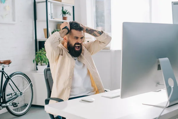 Empresário barbudo impulsivo com as mãos na cabeça sentado na mesa do computador e gritando no escritório — Fotografia de Stock