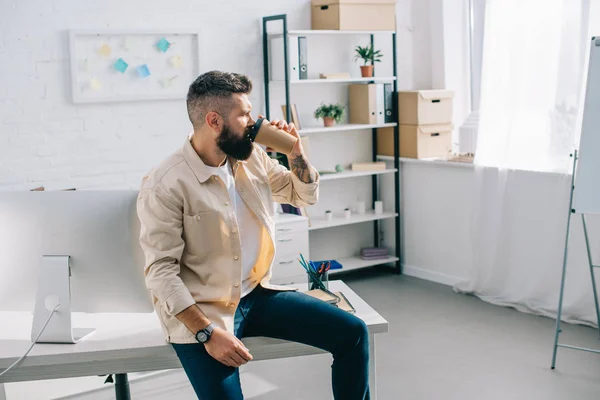 Bearded businessman sitting on office desk and drinking coffee from disposable cup — Stock Photo