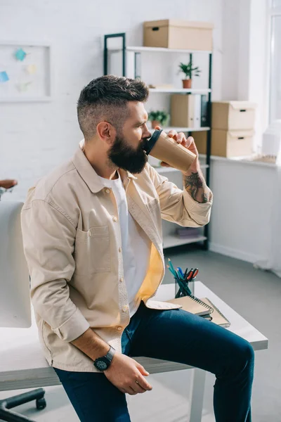Бородатий бізнесмен сидить на столі і п'є каву з паперової чашки в офісі — стокове фото