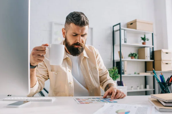 Male designer sitting with white cup of tea and looking at palette in office — Stock Photo