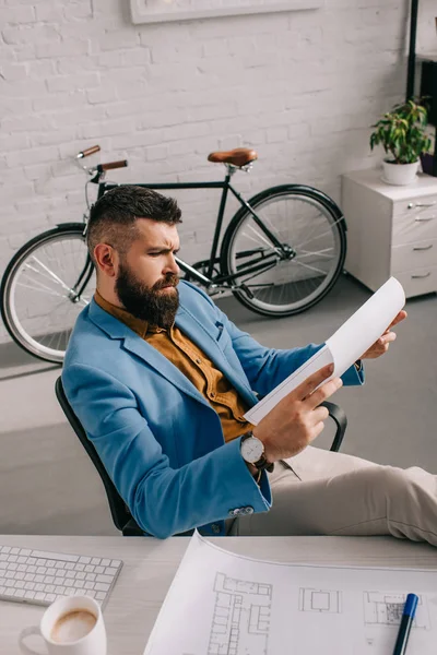 Мужчина-архитектор в синей куртке сидит и смотрит на бумагу в офисе — стоковое фото
