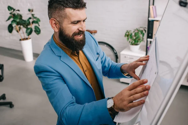 Arquiteto masculino olhando para o papel na placa branca e sorrindo no escritório moderno — Fotografia de Stock