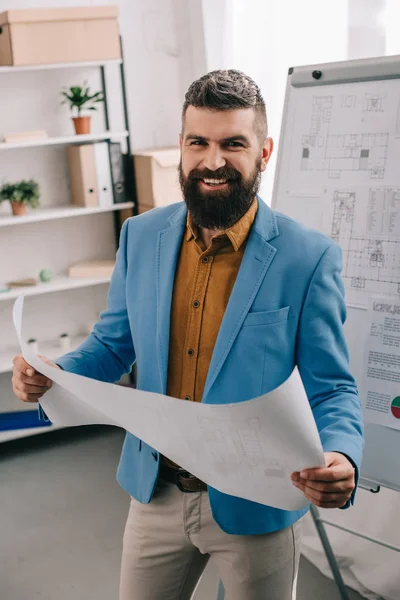 Schöner Architekt lächelt und hält Blaupause in modernem Büro — Stockfoto