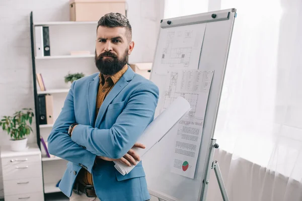 Serious adult male architect in blue formal wear looking at camera, holding blueprint, using flip chart and working on project in office — Stock Photo