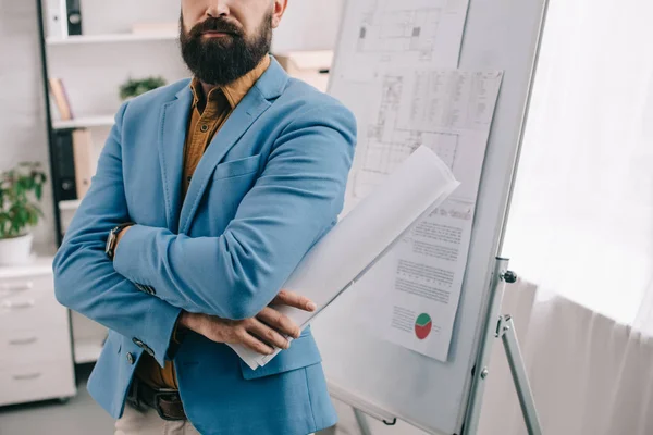 Vue recadrée de l'architecte masculin adulte en tenue formelle bleue tenant le plan, utilisant le tableau à feuilles mobiles et travaillant sur le projet dans le bureau — Photo de stock