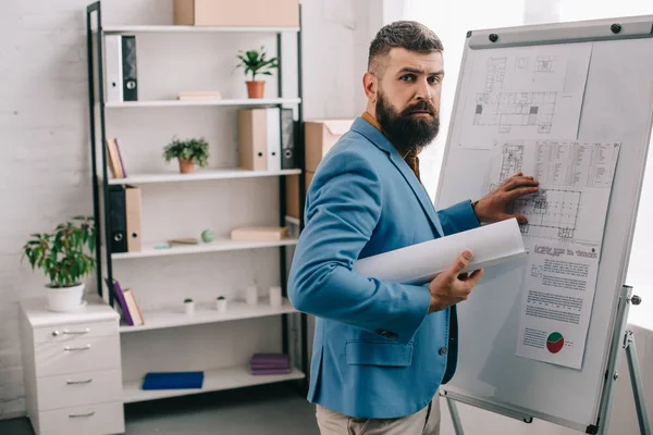 Bonito adulto masculino arquiteto no azul formal desgaste segurando blueprint, usando flip chart e trabalhando no projeto no escritório — Fotografia de Stock