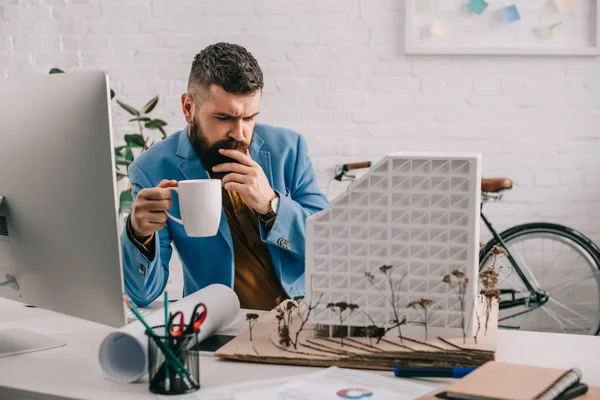 Arquitecto masculino adulto enfocado en ropa formal sosteniendo taza de café y trabajando en el proyecto en la oficina - foto de stock