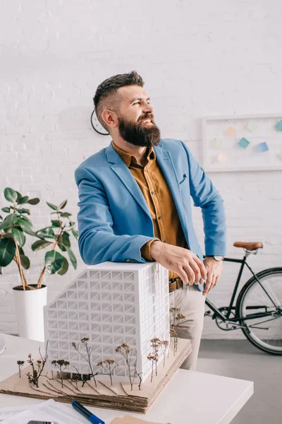 Happy adult male architect in formal wear standing near house model in office — Stock Photo