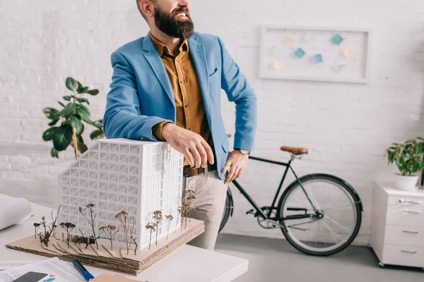 Ausgeschnittene Ansicht eines glücklichen erwachsenen männlichen Architekten in formeller Kleidung, der in der Nähe eines Hausmodells im Büro steht — Stockfoto