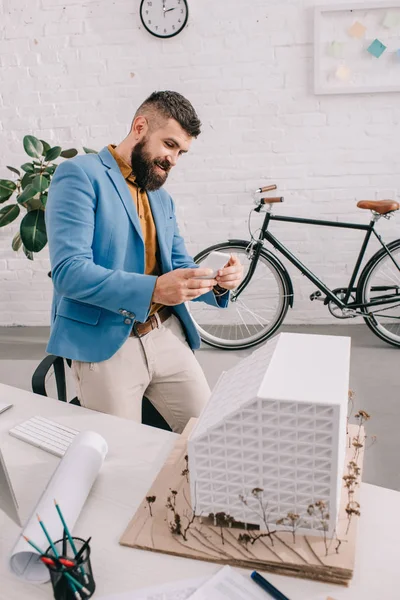 Sorridente adulto arquiteto masculino em desgaste formal tirar foto no smartphone do modelo de casa no escritório — Fotografia de Stock