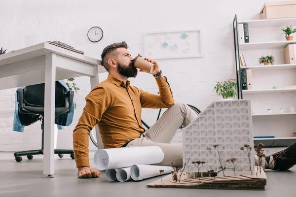 Barbudo adulto arquiteto masculino beber café para ir e trabalhar em projeto de construção com plantas no escritório — Fotografia de Stock
