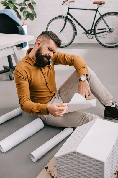 Concentrato barbuto adulto maschio architetto seduto sul pavimento e lavorando su cianografie in ufficio — Foto stock