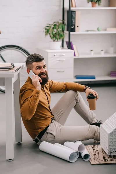 Architetto maschio adulto barbuto sorridente parlando su smartphone, tenendo il caffè per andare e lavorando su cianografie in ufficio — Foto stock