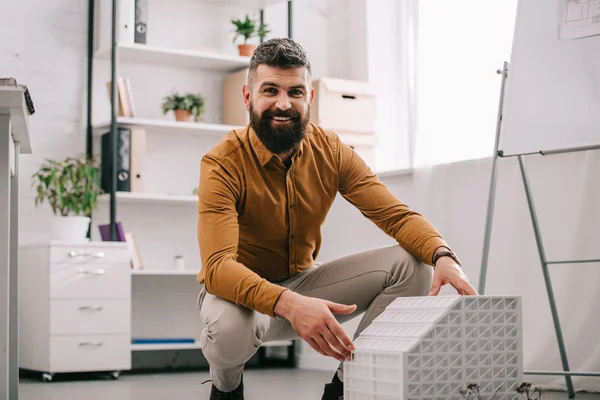 Sorridente barbudo adulto arquiteto masculino olhando para a câmera e apresentando modelo de casa no escritório — Fotografia de Stock