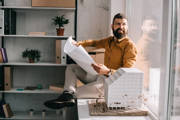 Sorridente barbudo adulto arquiteto masculino sentado perto de casa modelo, segurando planta e trabalhando no projeto no escritório — Fotografia de Stock