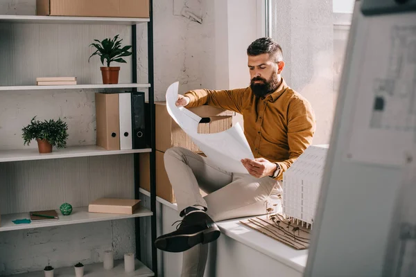 Arquiteto masculino adulto barbudo pensivo sentado perto do modelo da casa, segurando o modelo e trabalhando no projeto no escritório — Fotografia de Stock