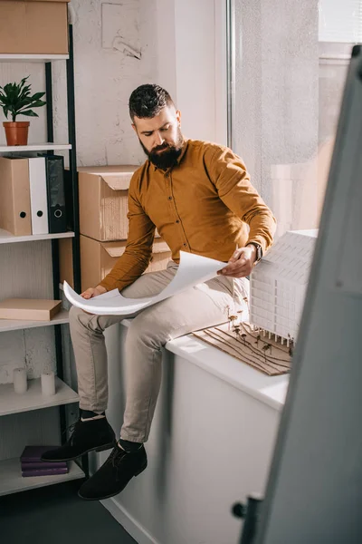 Задумчивый бородатый взрослый мужчина-архитектор сидит рядом с моделью дома, держит чертеж и работает над проектом в офисе — стоковое фото