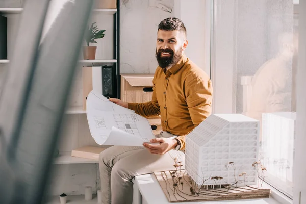 Architetto maschio adulto barbuto sorridente seduto vicino al modello di casa, che tiene il progetto e lavora al progetto in ufficio — Foto stock