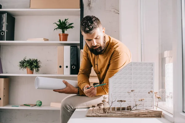 Ориентированный бородатый взрослый архитектор мужского пола, держащий чертеж, указывающий на модель дома и работающий над проектом в офисе — стоковое фото