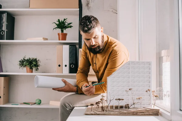 Architecte masculin adulte barbu concentré tenant le plan directeur, pointant vers le modèle de la maison et travaillant sur le projet dans le bureau — Photo de stock