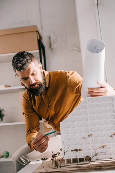 Arquitecto masculino adulto barbudo enfocado sosteniendo el plano, apuntando al modelo de casa y trabajando en el proyecto en la oficina - foto de stock