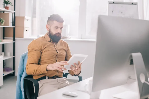 Hombre de negocios adulto enfocado sentado en el escritorio de la oficina y usando tableta digital en el lugar de trabajo - foto de stock