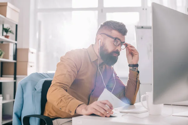 Serio hombre de negocios adulto barbudo en auriculares sentado y trabajando en el escritorio de la computadora en la oficina - foto de stock