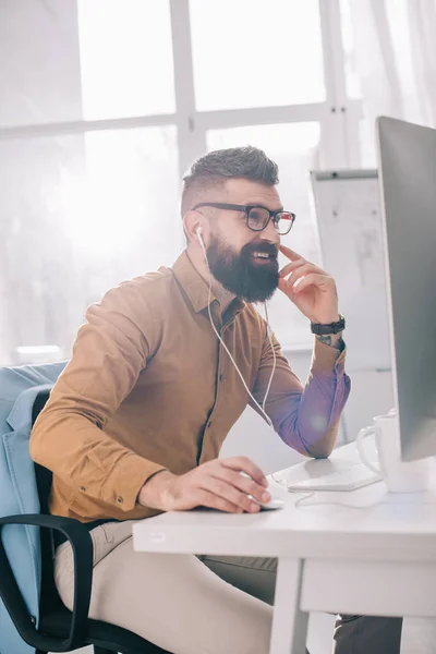 Lächelnder bärtiger erwachsener Geschäftsmann mit Kopfhörern, der im Büro am Computertisch sitzt und arbeitet — Stockfoto