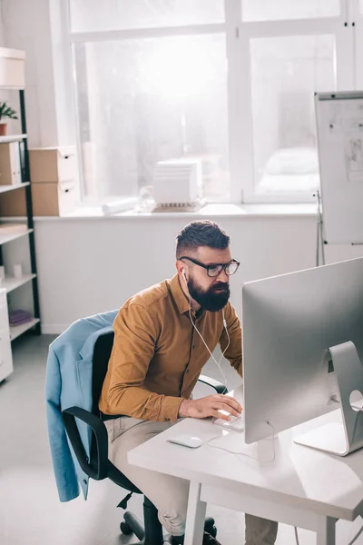Бородатий дорослий бізнесмен в навушниках сидить і працює за комп'ютерним столом в офісі — стокове фото