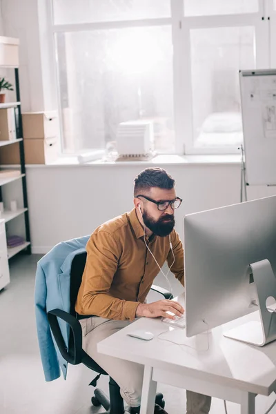 Bearded adult businessman in earphones sitting and working at computer desk in office — Stock Photo