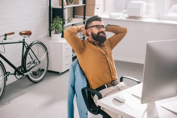 Smiling bearded adult businessman in earphones with hands behind head sitting and relaxing at computer desk in office — Stock Photo