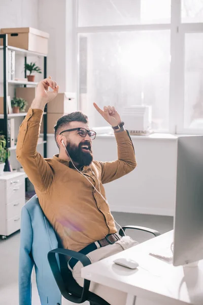 Happy bearded adult businessman in earphones sitting at computer and listening to music desk in office — Stock Photo