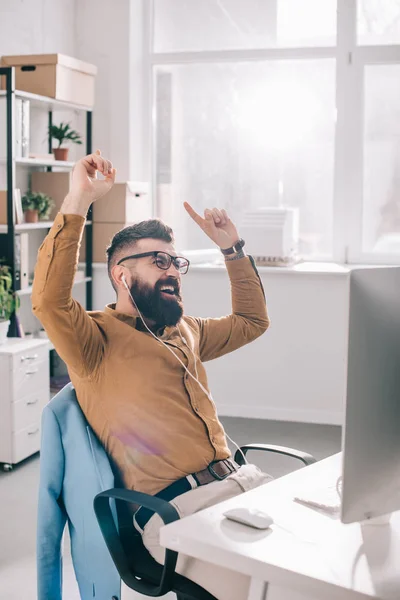 Heureux homme d'affaires adulte barbu dans les écouteurs assis à l'ordinateur et à l'écoute du bureau de musique dans le bureau — Photo de stock