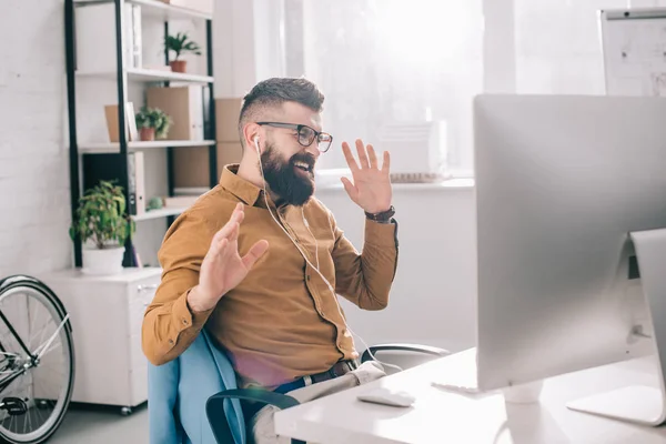 Excited bearded adult businessman in earphones sitting at computer and listening to music desk in office — Stock Photo