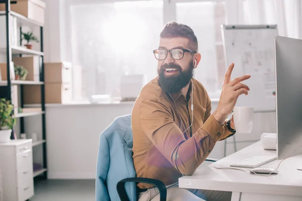 Sonriente hombre de negocios adulto barbudo en auriculares sentado y trabajando en el escritorio de la computadora en la oficina - foto de stock