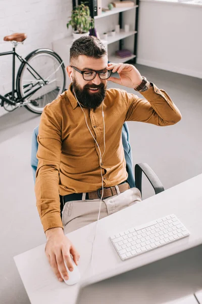 Lächelnder bärtiger erwachsener Geschäftsmann mit Kopfhörern, der im Büro am Computertisch sitzt und arbeitet — Stockfoto