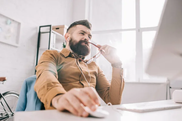 Pensive bearded adult businessman in earphones sitting and working at computer desk in office — Stock Photo