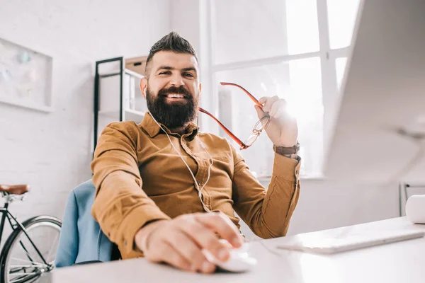 Усміхнений бородатий дорослий бізнесмен в навушниках дивиться на камеру і працює за комп'ютерним столом в офісі — стокове фото