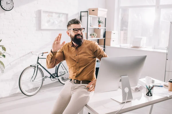 Cheerful bearded adult businessman sitting at office desk, looking away and waving — Stock Photo