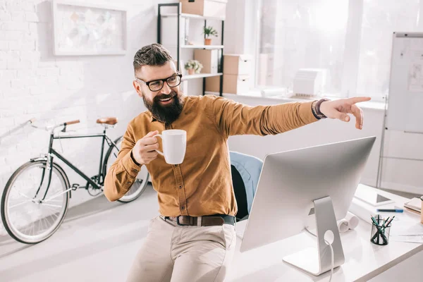 Homem de negócios adulto barbudo feliz segurando xícara de café e apontando o dedo na saída no local de trabalho — Fotografia de Stock