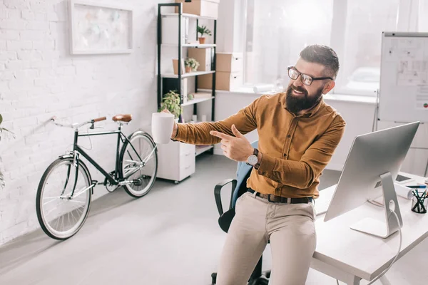 Feliz barbudo adulto hombre de negocios sosteniendo taza de café y señalando el dedo en la salida en el lugar de trabajo - foto de stock