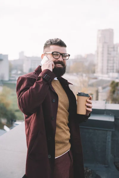 Hombre adulto con estilo en gafas hablando en el teléfono inteligente y la celebración de café para ir en la azotea - foto de stock