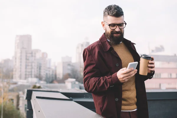 Homem adulto elegante em óculos segurando café para ir e usando smartphone no telhado — Fotografia de Stock